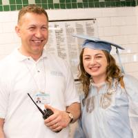 A man wearing a white polo shirt holds a radio and poses next to a graduate wearing a CU cap and gown
